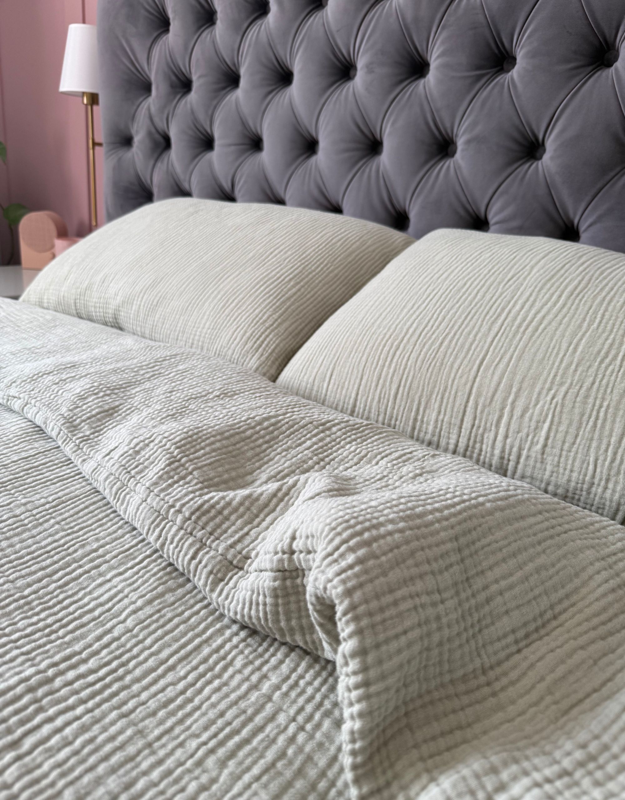 A close up of light green muslin bedding and two pillows on a bed with a dark blue-gray headboard and pink-painted wall-paneling behind