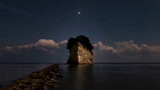 A bright point of light — Venus — appears in the dark sky above a large rock structure surrounded by water.