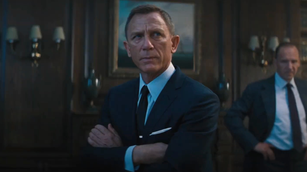 Daniel Craig está sentado com os braços cruzados parecendo irritado enquanto Ralph Fiennes assiste No Time To Die.