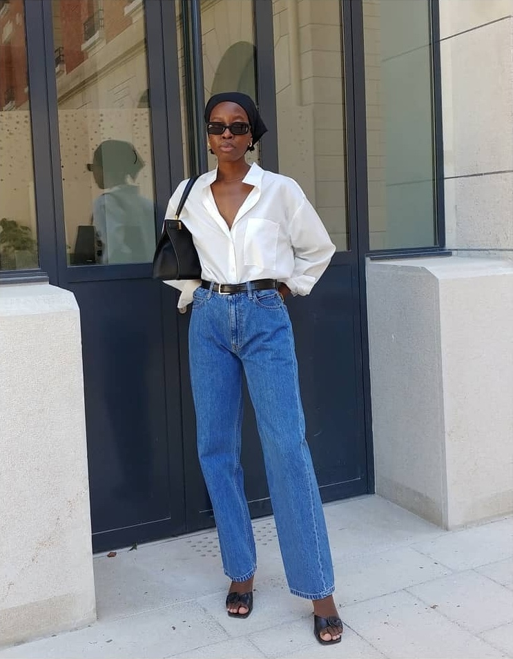 style influencer Sylvie Mus poses on a Paris sidewalk wearing a white button-down shirt, black leather shoulder bag, black headscarf, belt, straight-leg jeans, and mule kitten-heel sandals