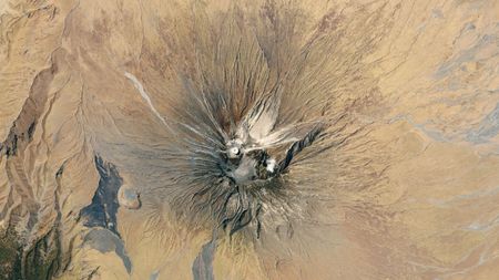 A satellite photo of a volcano surrounded by barren plains. Light colored lava flows can be seen on its flanks and a bright white cone at its summit