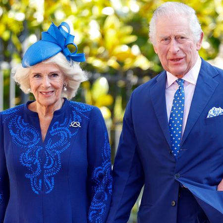 Queen Camilla wearing a royal blue dress and matching hat smiling with King Charles who is wearing a blue suit and a blue and white polka dot tie