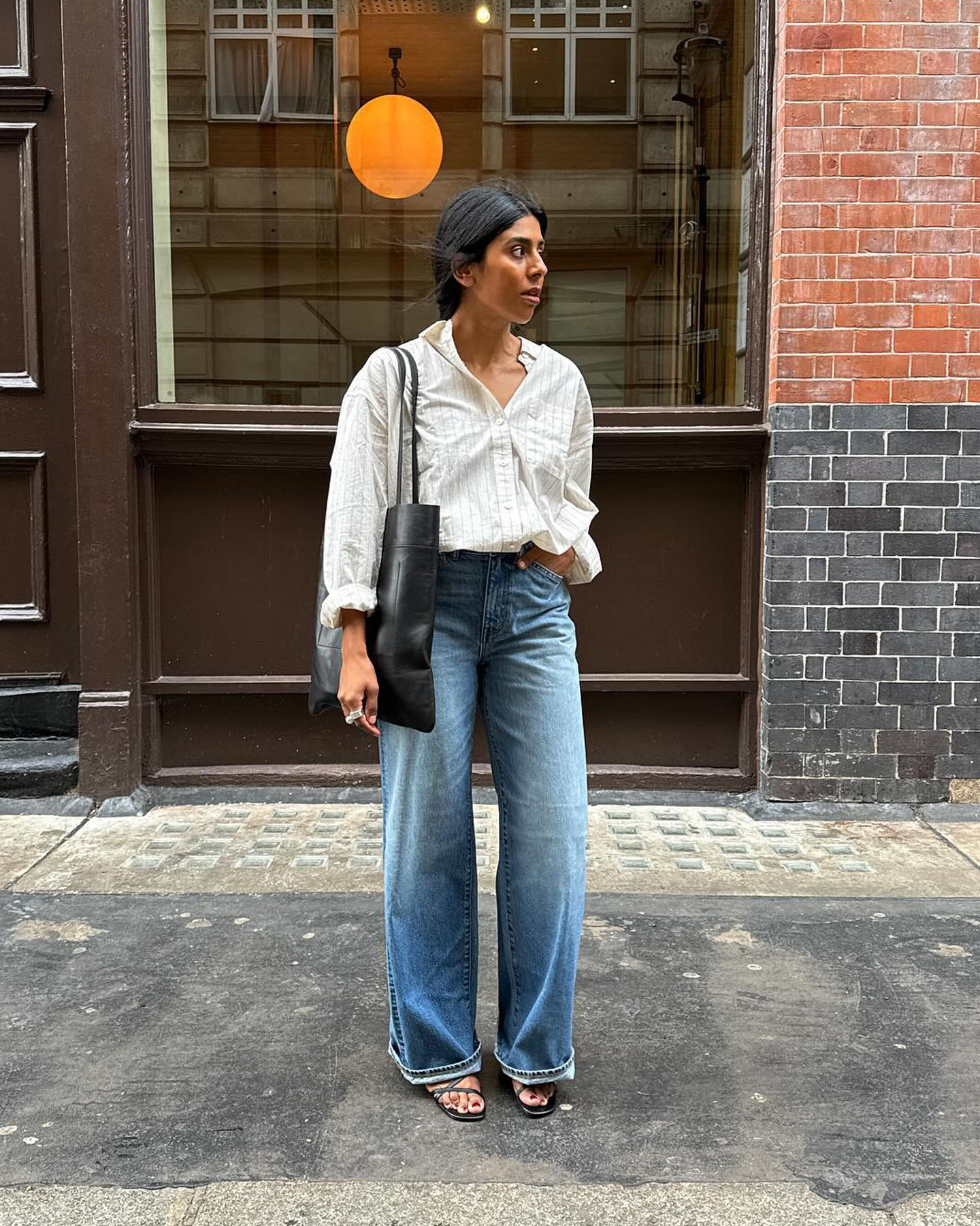 British fashion influencer Monikh poses on the sidewalk wearing a striped white button-down shirt, cuffed jeans, and strappy sandals