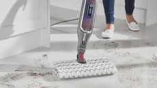 A Shark S6003UK Klik n Flip Automatic Steam Mop Cleaner being used on muddy white tiled flooring by female wearing denim jeans and casual white leather flat loafer-style footwear