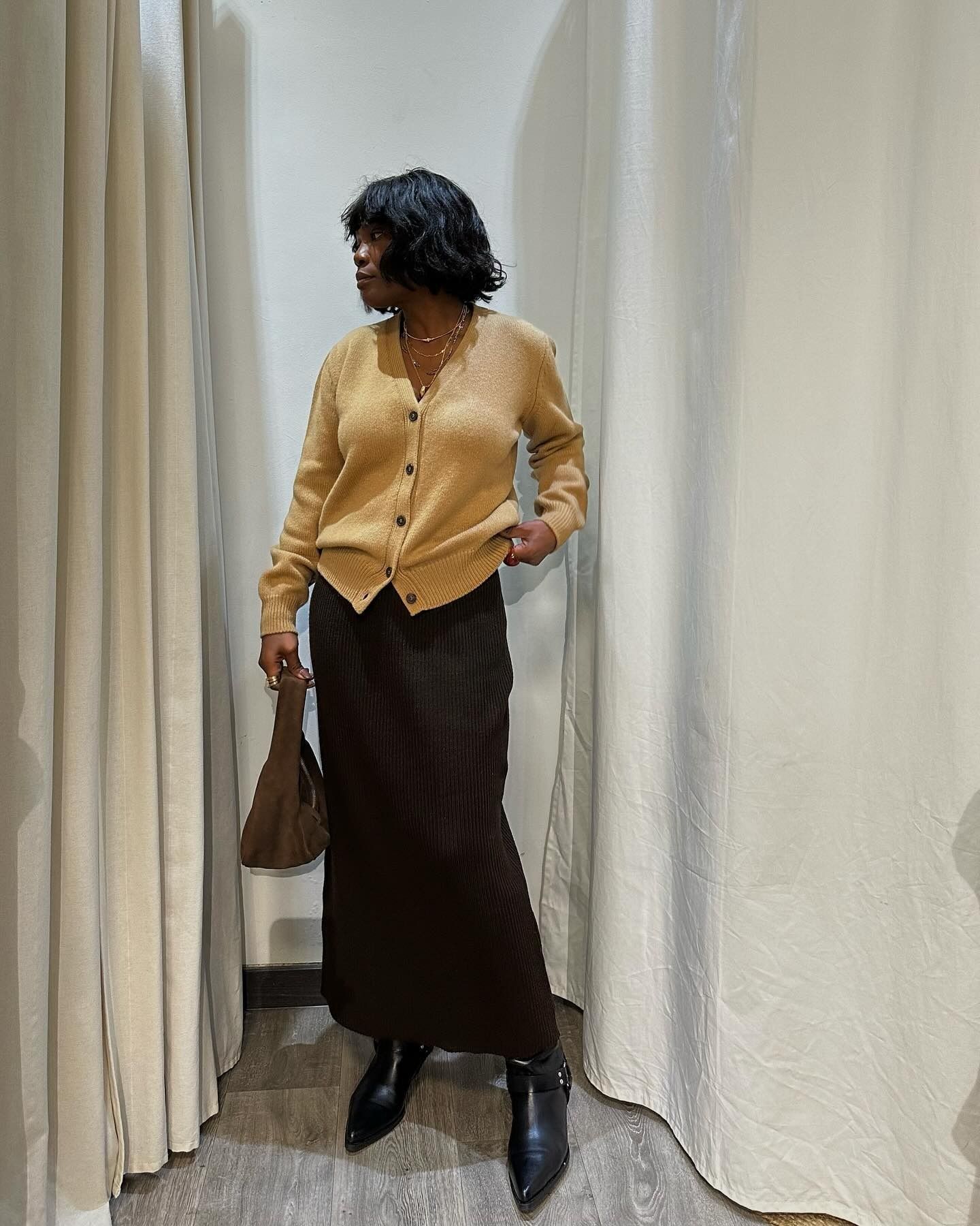 Style influencer Franny Mozemba poses in a camel button-front cardigan, brown suede bag, black maxi skip skirt, and black pointed-toe boots.