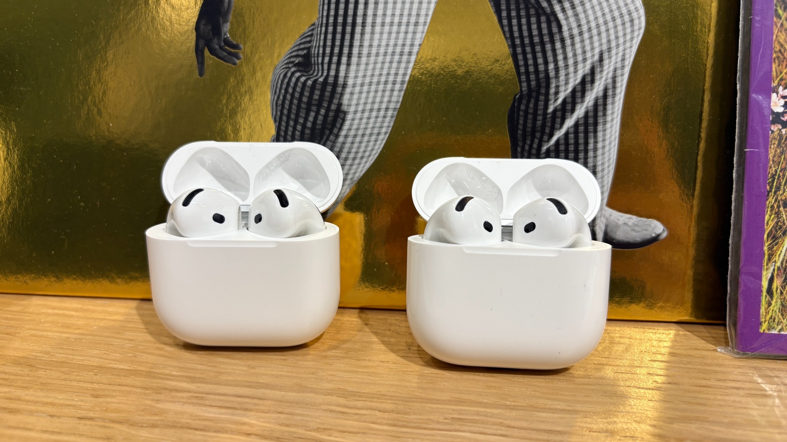The AirPods 5 and AirPods 4 in their charging cases next to each other on a wooden surface.