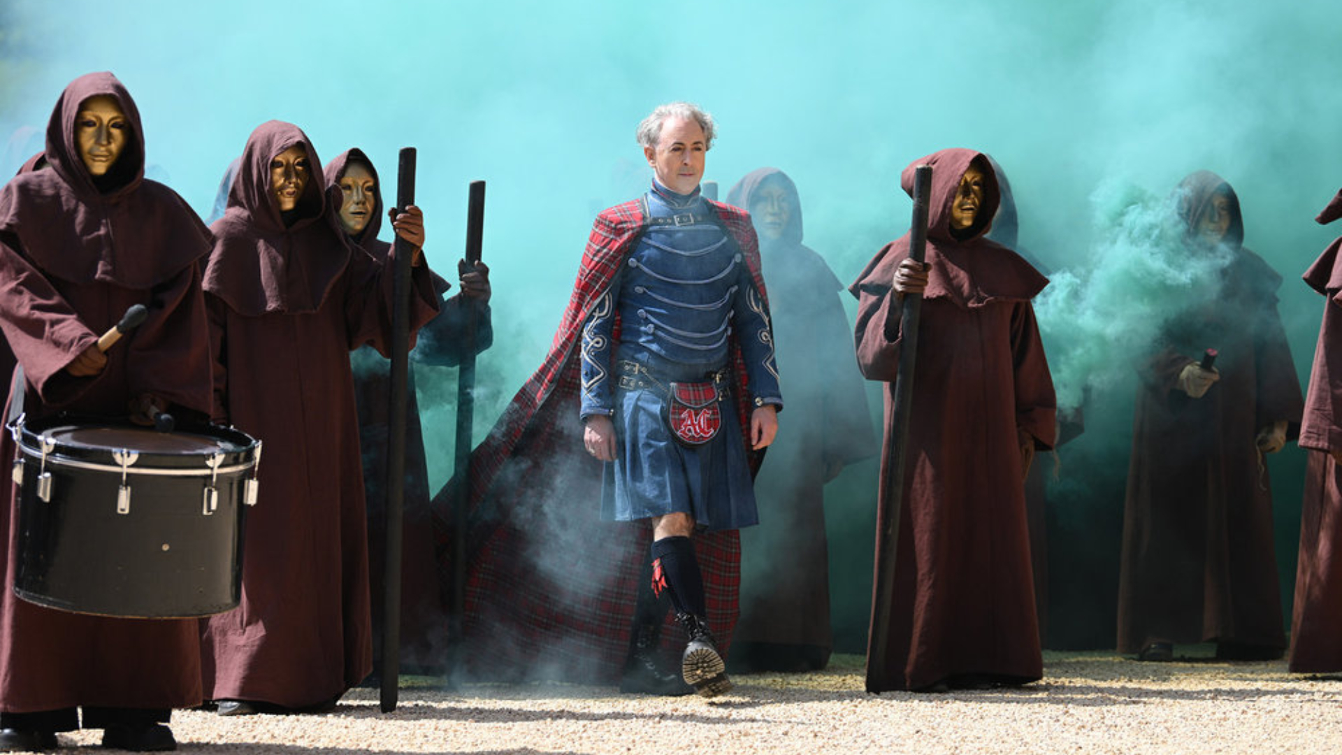 Host Alan Cumming wears armor and a cape while standing amid cloaked figures with gold masks in a cloud of blue smoke, in NBC's 'The Traitors: New Blood.'