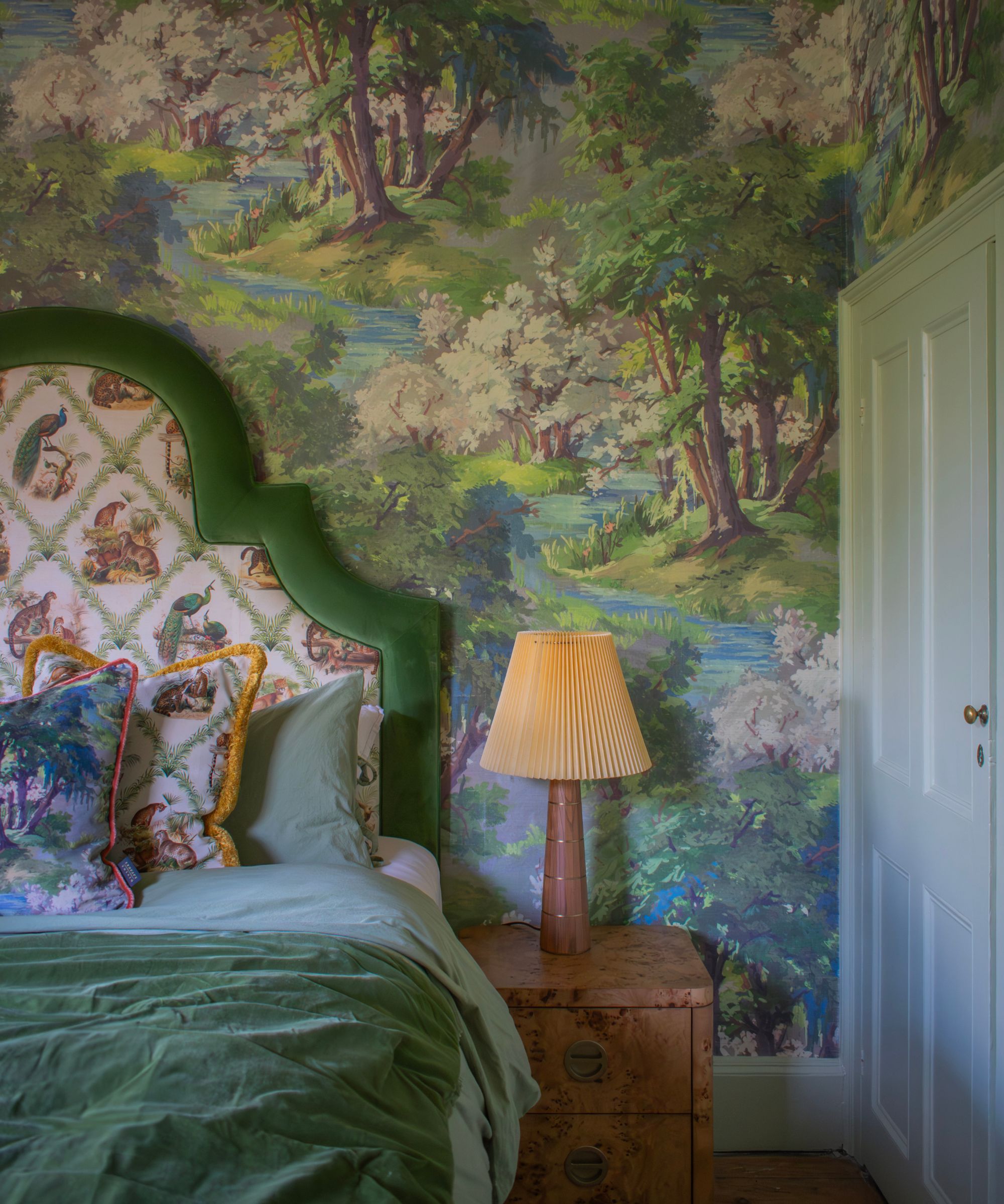 A bedroom featuring a painted landscape mural of trees, water and greenery covering the wall. A green upholstered headboard frames the bed. Soft bedding, patterned cushions and a bedside lamp create a calm, storybook-like sleeping space with gentle colour harmony.
