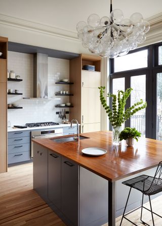 kitchen with blond wood cabinetry, white backsplash, grey units, grey kitchen island with wooden countertop, glass bubble chandelier, wooden floors, open shelving