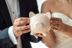 A heterosexual couple in a suit and wedding gown hold a piggy bank together.