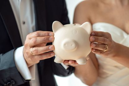 A heterosexual couple in a suit and wedding gown hold a piggy bank together.
