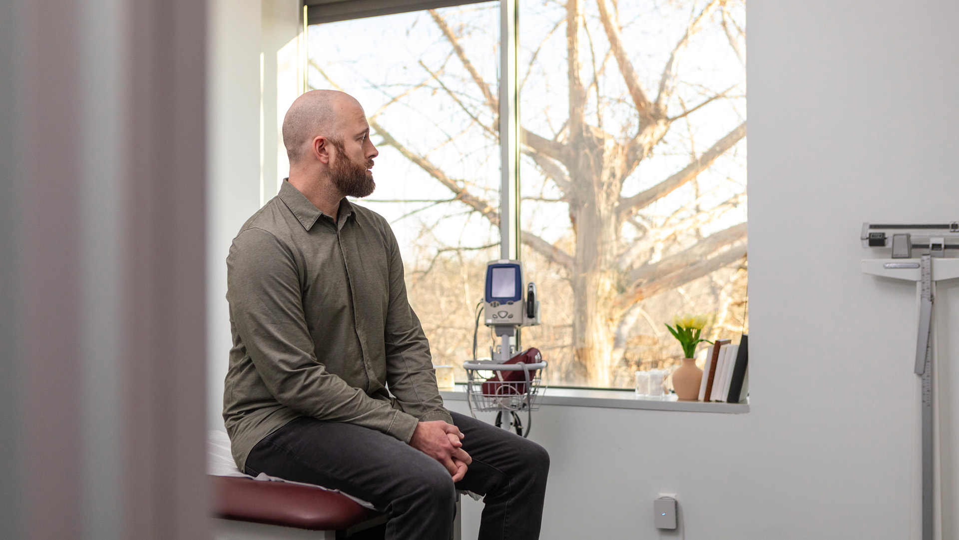 A bald man in a green shirt with long sleeves and dark pants sits on a hospital bench with medical equipment next to them.