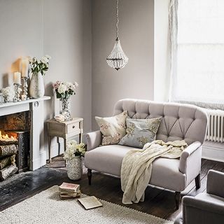 light grey living room with wooden floor, with a fire burning in the fireplace to the left and an elegant light purple sofa with cushions and a throw draped over it