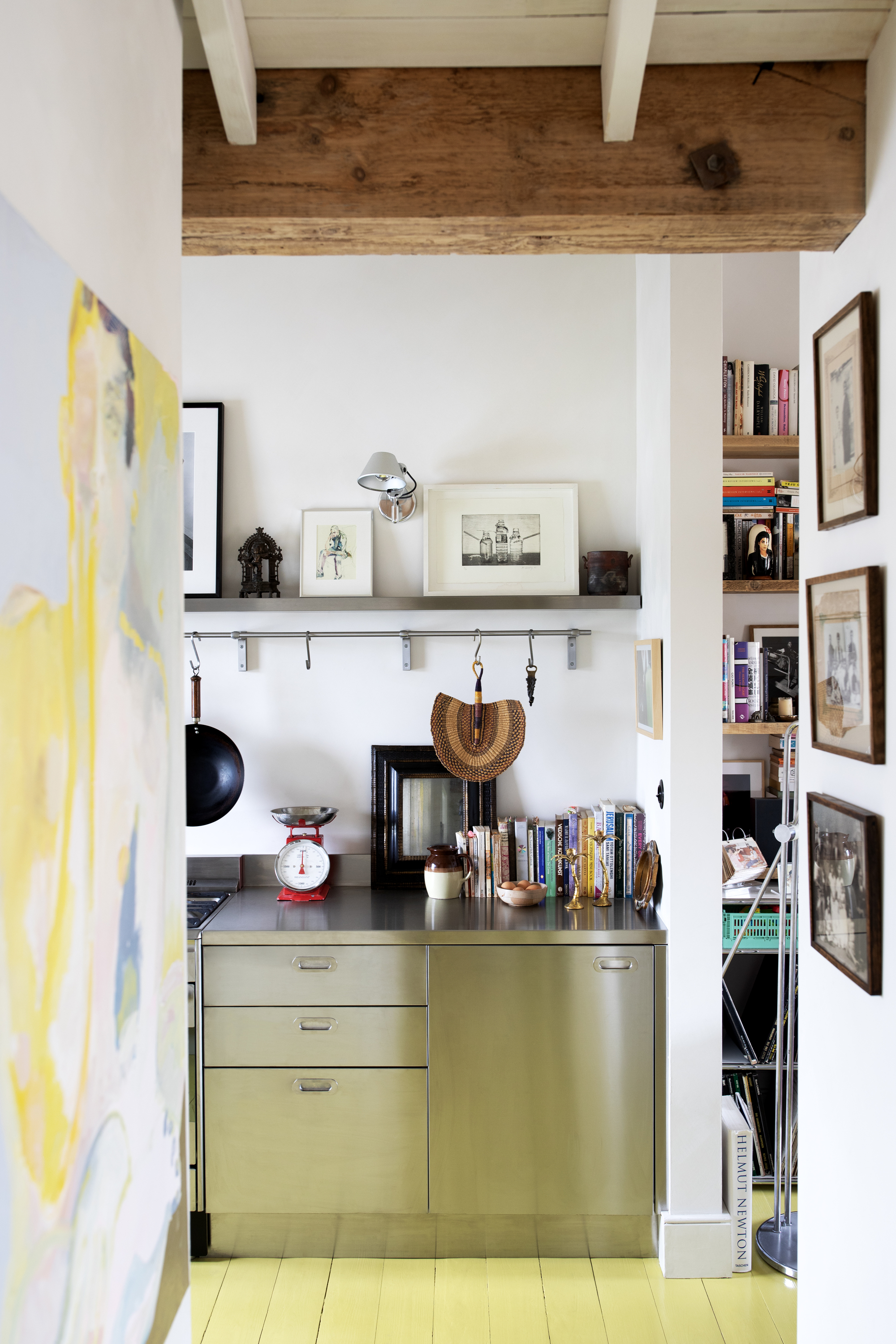 small stainless steel kitchen with white walls and open shelf, cookbooks styled on the counter, large artwork, timber ceiling beam, and yellow painted floorboards