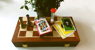 Bästa sällskapsspel: Ett gäng spelkort och ett timglas står på ett schackbräde bredvid två schackpjäser på ett vitt bord, med en grön krukväxt bakom.