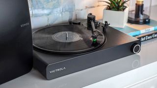 The Victrola Stream Onyx turntable pictured on.a white shelf next to a Sonos speaker on one side and pile of books on the other.
