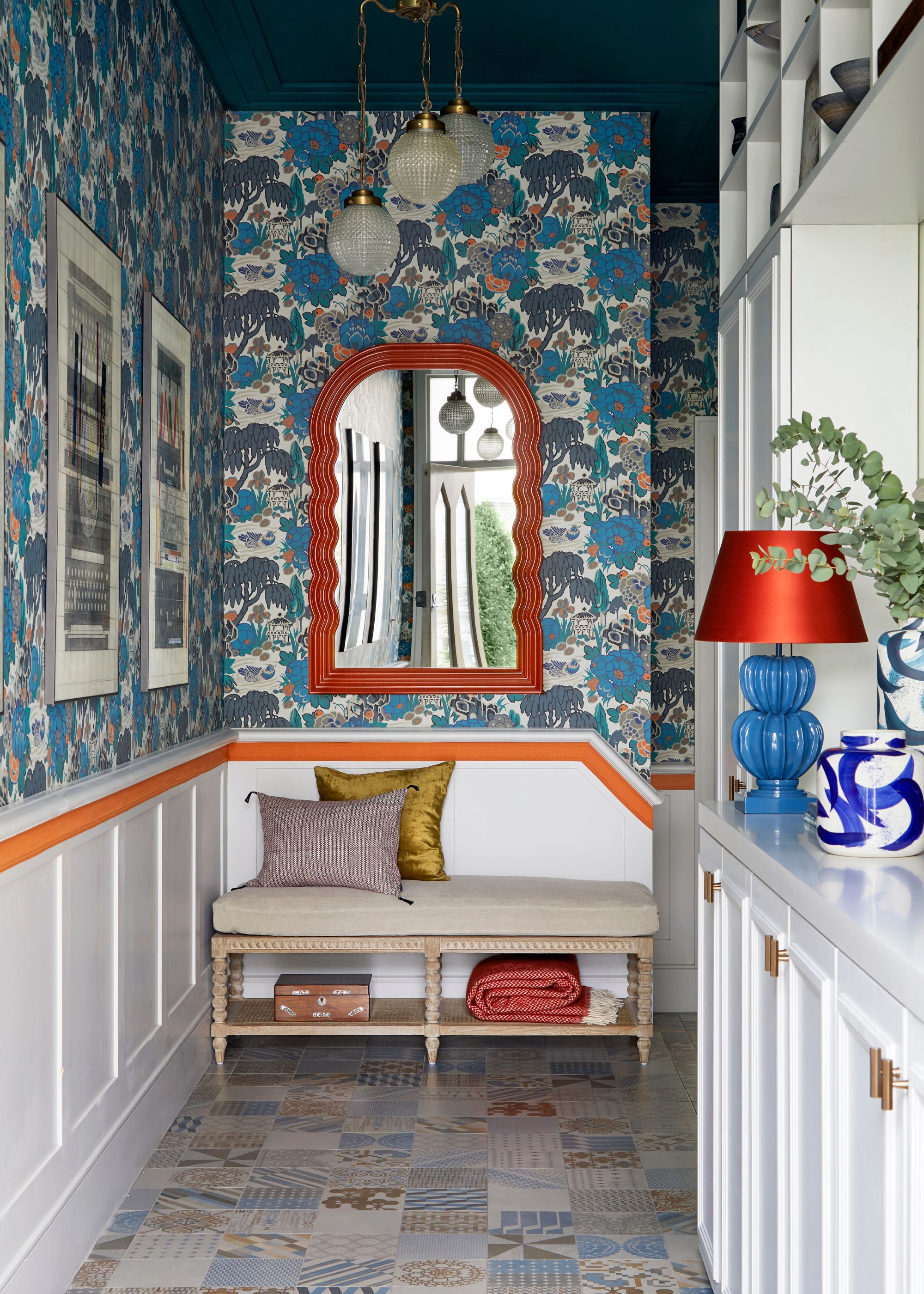 entryway with blue patterned wallpaper, bright orange wall mirror, bench with cushions and storage underneath