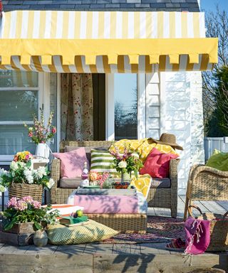 An example of patio cover ideas showing a yellow and white striped canopy with colourful textiles on wicker garden furniture