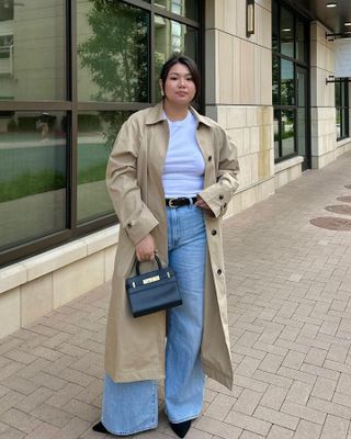 female fashion influencer Marina Torres poses on a sidewalk wearing a stylish outfit with a trench coat, white tee, wide leg jeans, and pointed-toe shoes