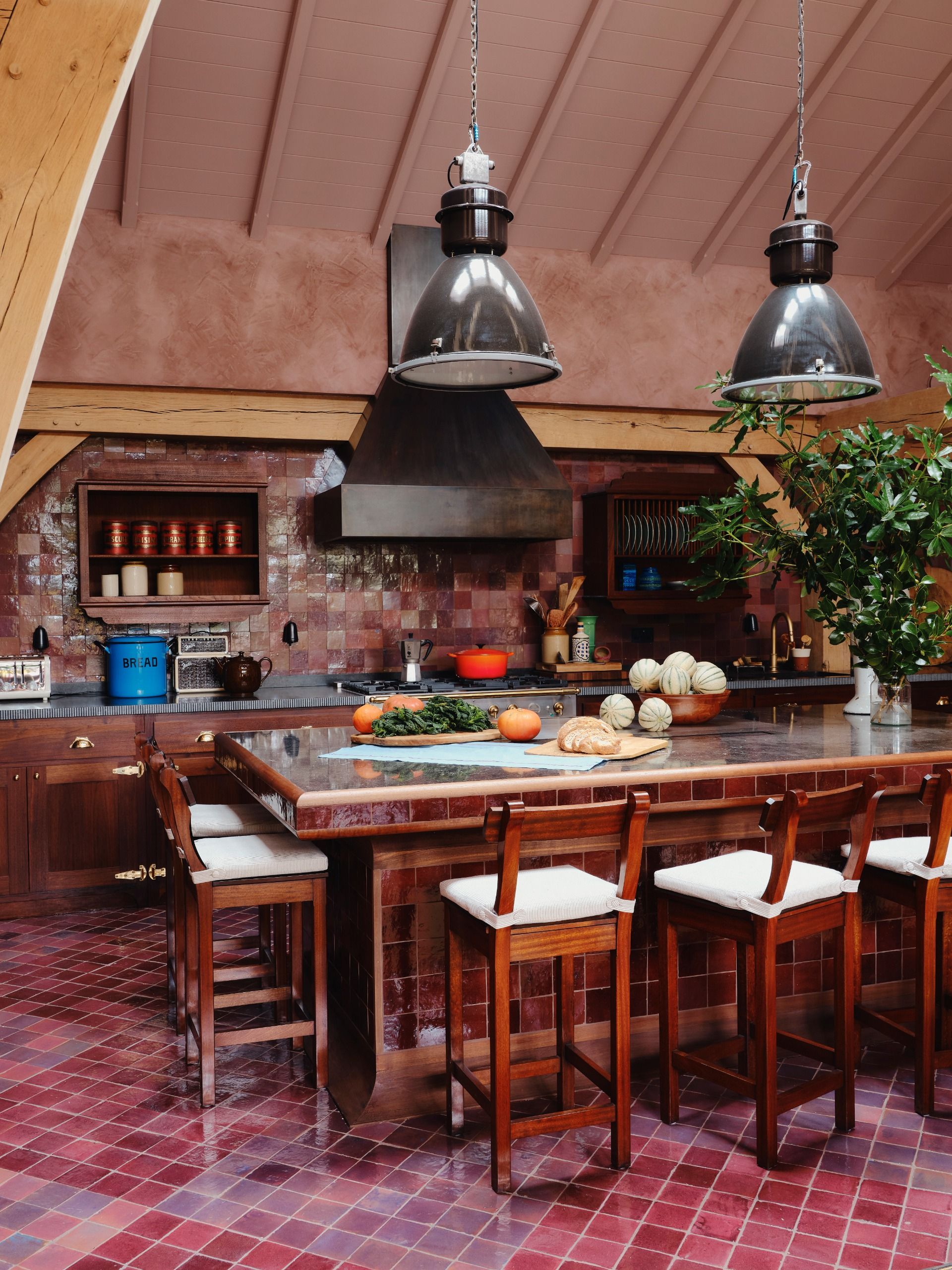A kitchen with a central island tiled, like its walls and floors, in tones of earth red, burgundy, and brown, decorated with wooden stools, white cushions, industrial style lighting, and plants.