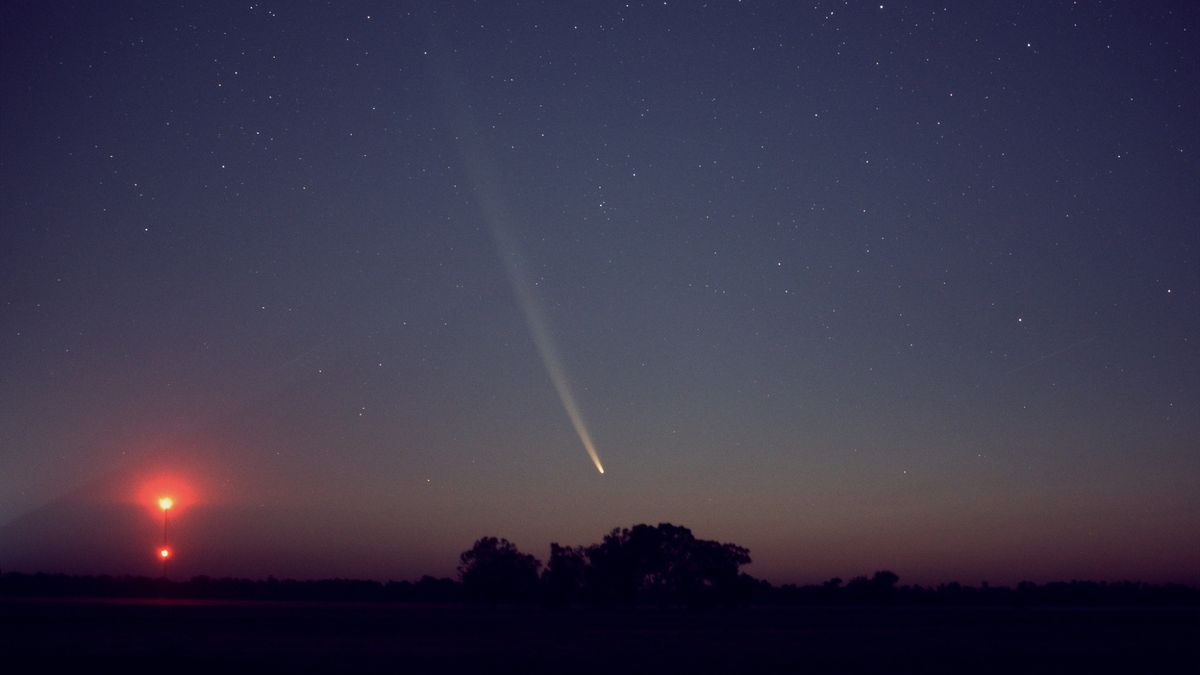 A comet streaks through a dim night sky as a faint sun sets.