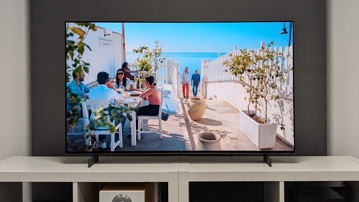 The LG C5 42-inch OLED TV seen straight-on on a TV cabinet. On screen are diners enjoying a restaurant's outdoor seating area with the sea in the background.