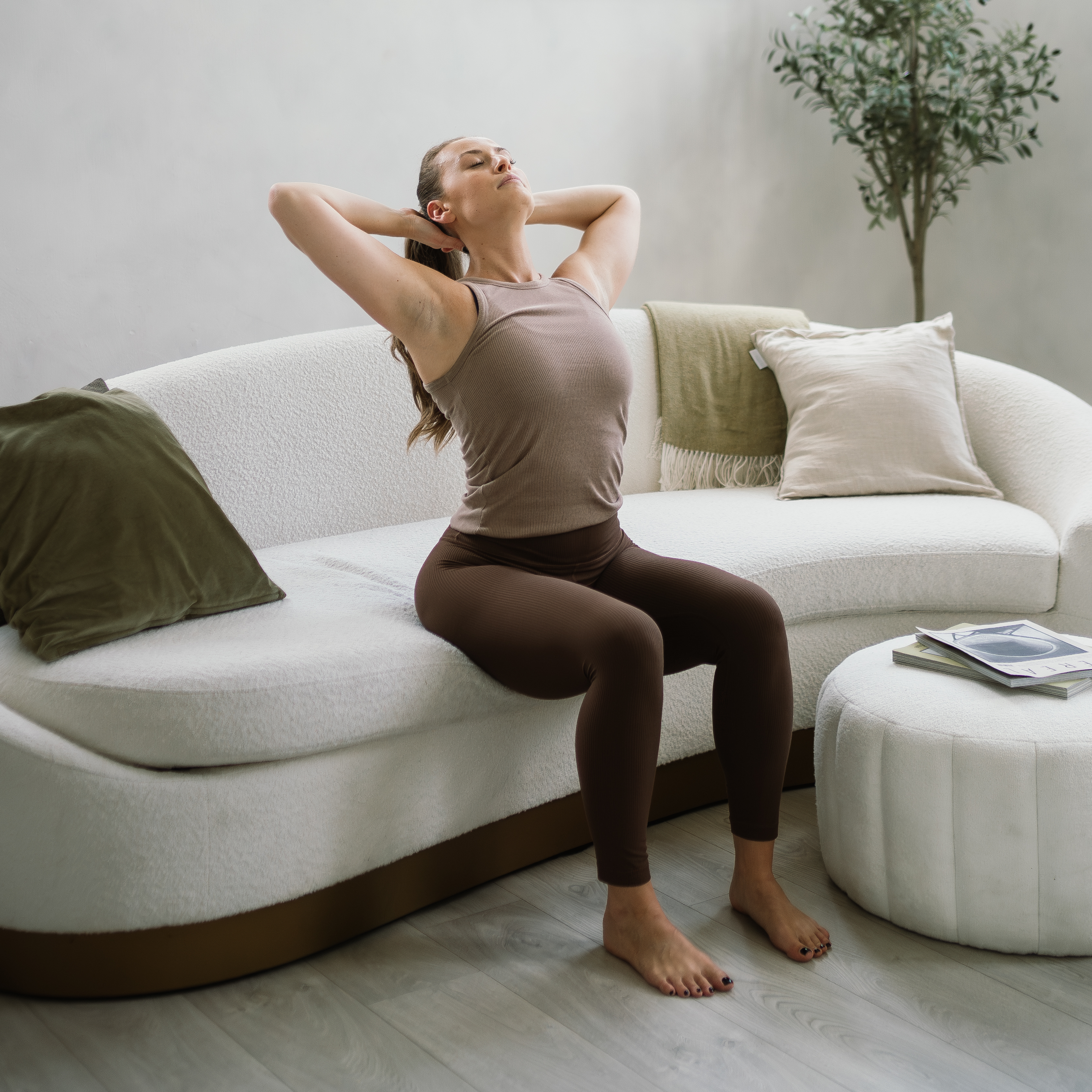 A woman sits on the edge of a curved white couch performing a spinal extension stretch. Her knees are bent, feet flat on the floor, buttocks on the edge of the couch, and her hands are behind her head as she stretches out her elbows and looks up to the ceiling.