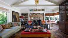Living room with white beamed ceiling, large persian rug, red ottoman, pale blue sofa with colourful cushions, blue striped armchairs and stone fireplace