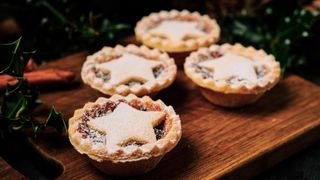 Four Mince pies dusted with icing sugar on a dark wood board with Holly and Pine cones