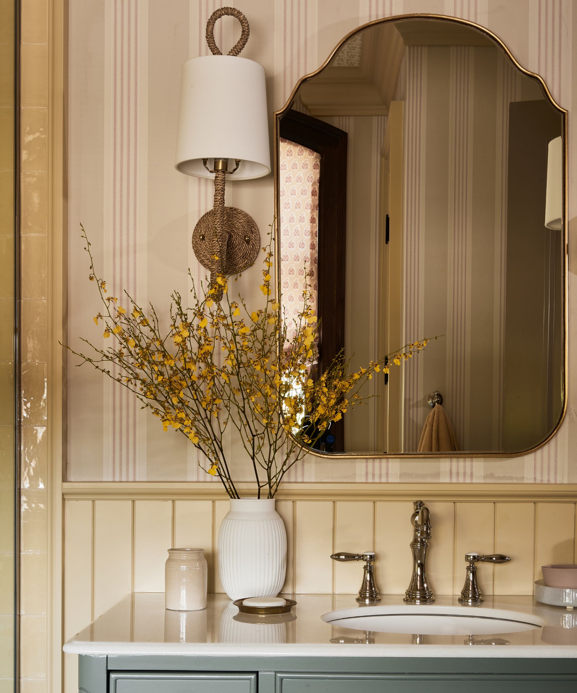 Close up of bathroom vanity area, yellow panelled walls, large mirror with wall light to the left, counterop with sink, vase and jars on top