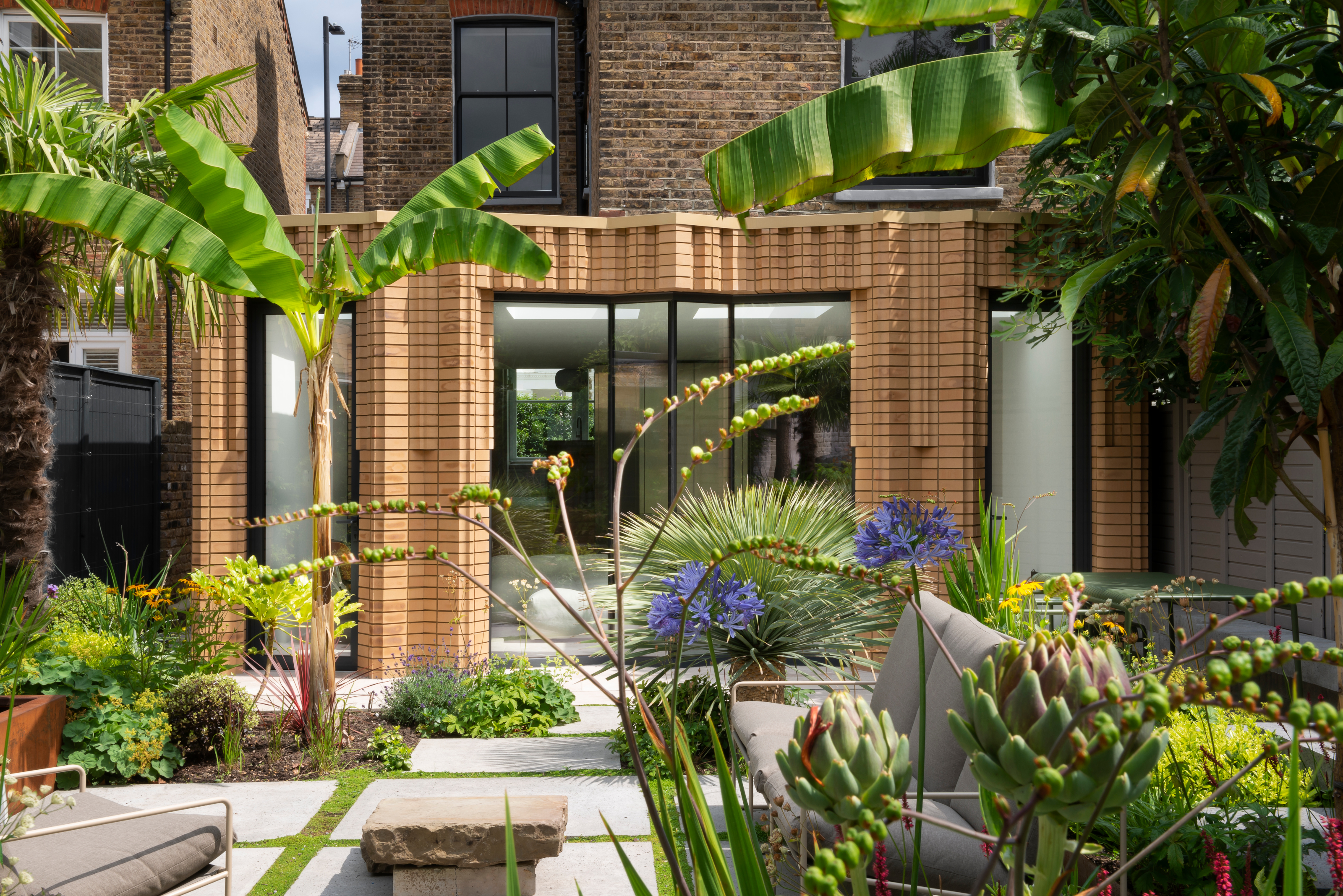 An extension with intricate brickwork, surrounding black-framed windows and doors leading out onto a tropical garden with colorful plants and banana leaf trees