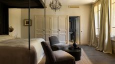 A bedroom with a pair of dark gray chairs, a black side table, a chandelier, and a four-poster bed