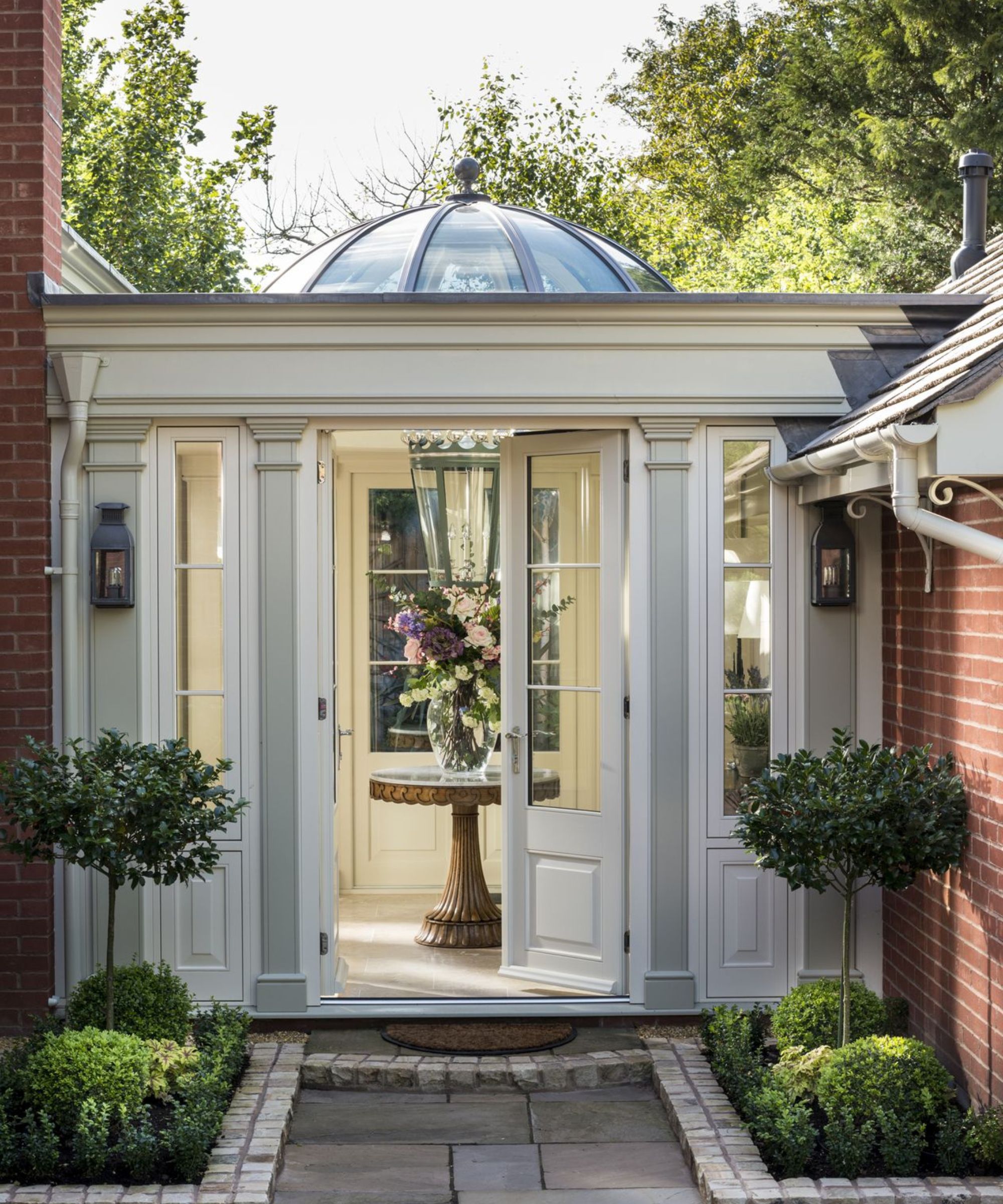 An elegant cream orangery porch with a glass dome roof, pilasters, French doors open to reveal a pedestal table with a large floral arrangement and clipped topiary either side.