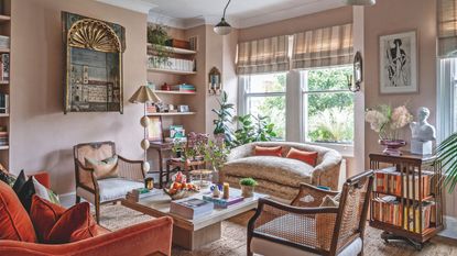 A warm, eclectic living room featuring dusty pink walls, a burnt orange velvet sofa, and woven cane armchairs arranged around a stone coffee table
