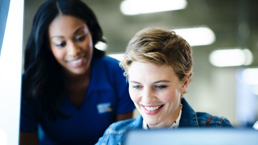 A smiling black woman in a blue uniform shirt leans in to assist a smiling white woman in a denim jacket who is looking at a tablet and a laptop screen.