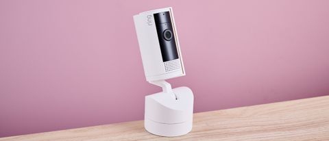 The Ring Pan-Tilt Indoor Cam sits on light-colored wood surface against a pink background.