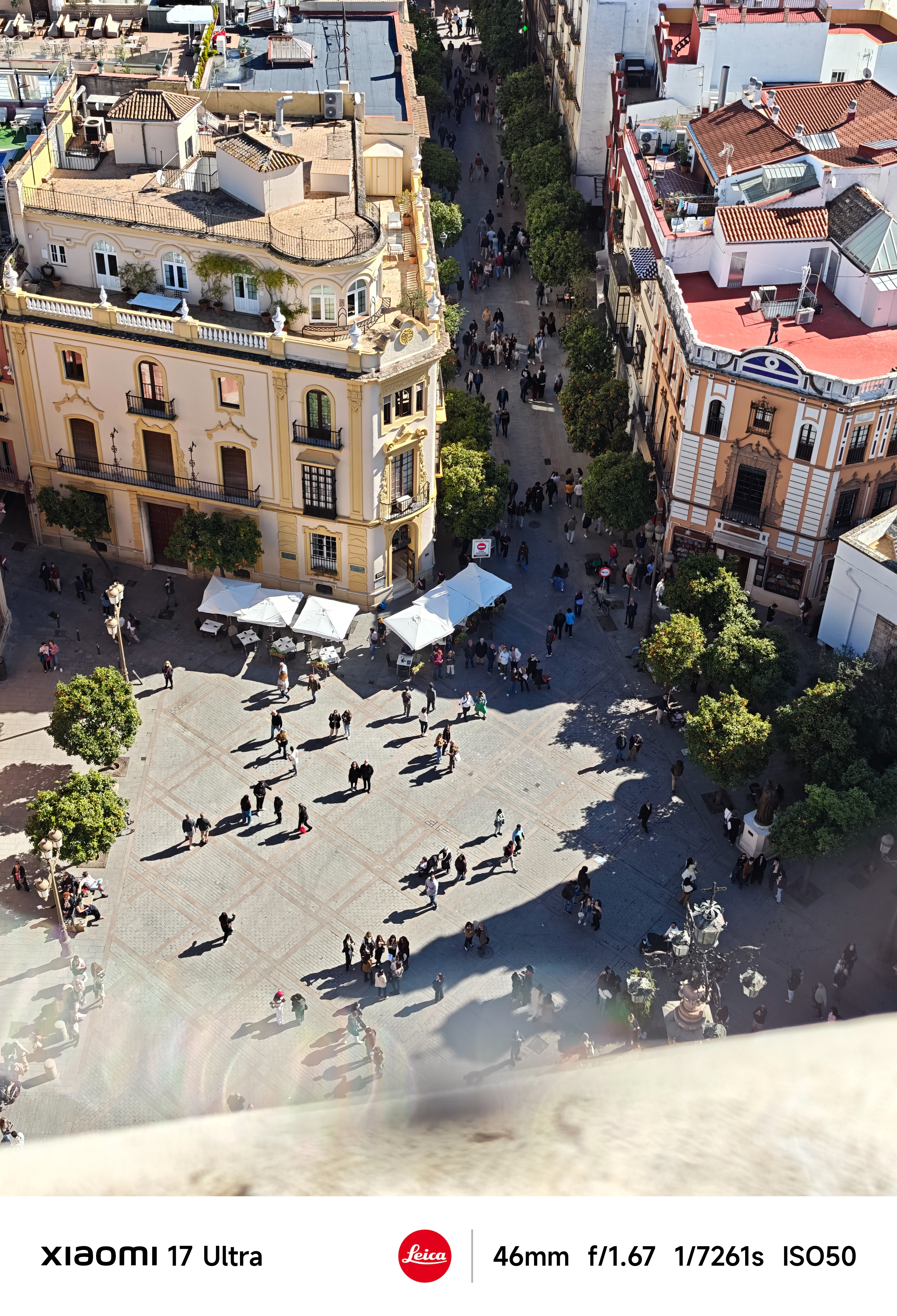 High-angle view of a busy plaza with people casting long shadows between historic buildings.
