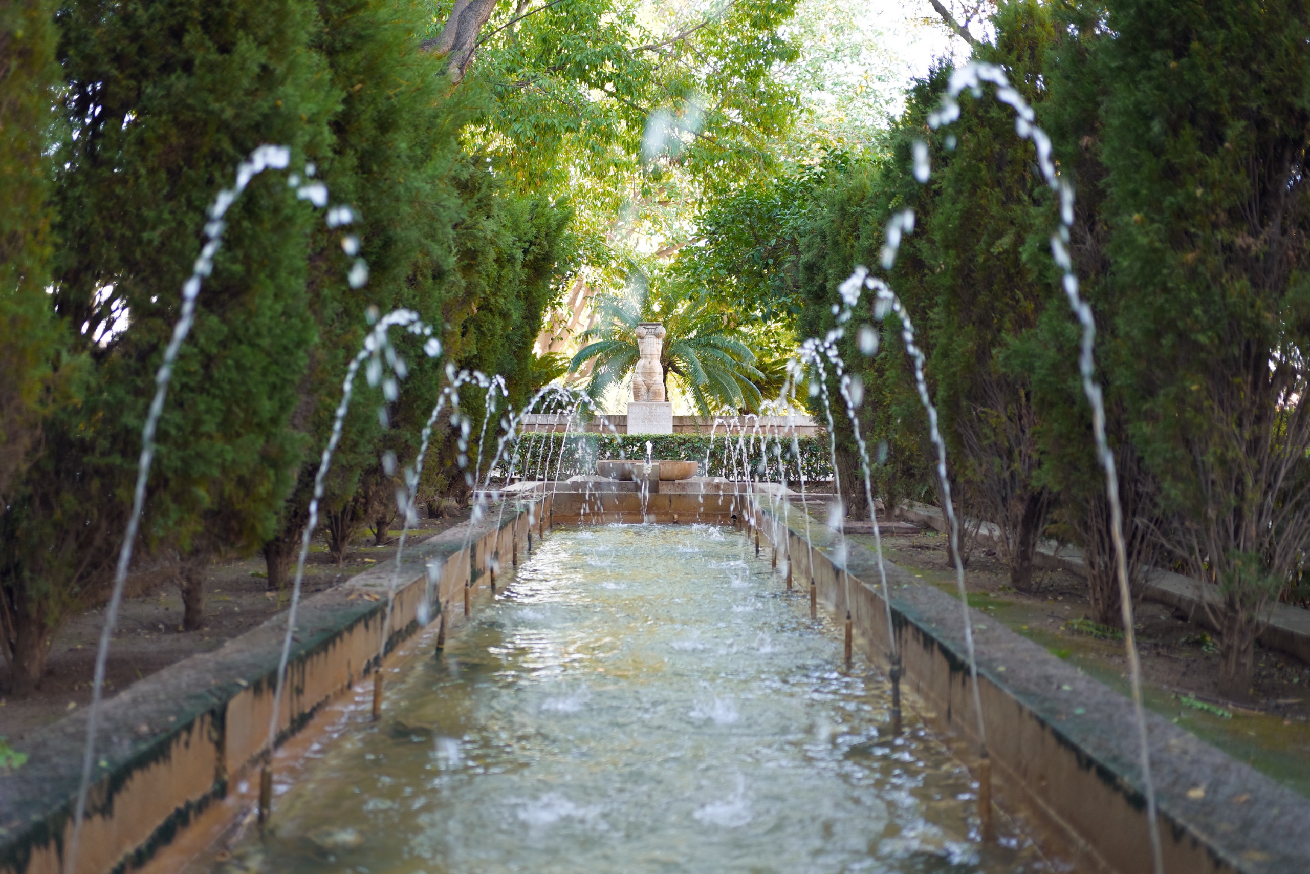 Long rectangular garden fountain with arcing water jets framed by dense green trees.