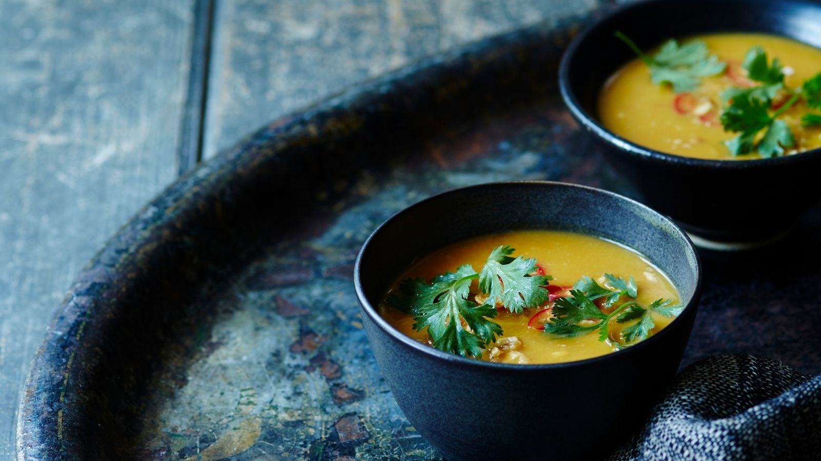 Two bowls of Thai butternut squash soup topped with herbs and chillies on a black tray