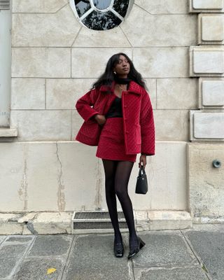 Woman wearing a red quilted jacket and matching skirt, nude-colored fleece-lined tights, and heels and standing in front of brick building.