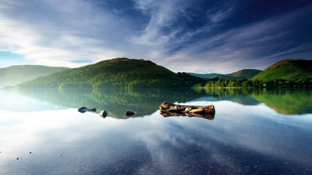 Serene lake surrounded by lush green hills under a blue sky with wispy clouds, featuring rocks and a log gracefully floating