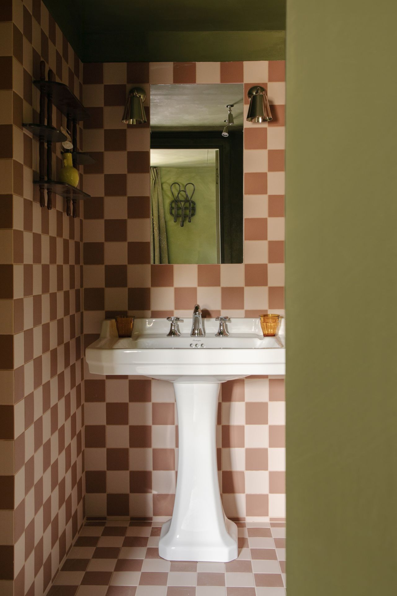 A checkered, terracotta and pale pink bathroom alternates patterned walls with olive green surfaces, brass accents, and a stuck-in-time atmosphere.