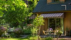 Exterior of a house with a garden with tall trees and an awning over a small patio are to suggest shadescaping ideas