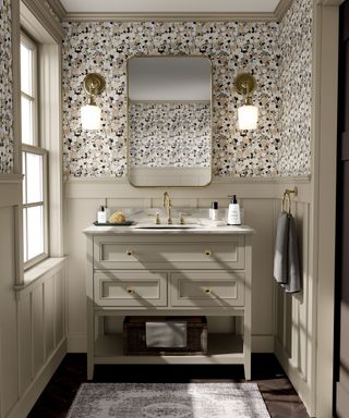 A small bathroom with beige and black dotted wallpaper, a large gold mirror with two wall sconces either side, a beige vanity unit with a gold faucet and soap dispensers, and a gray rug on the dark floor