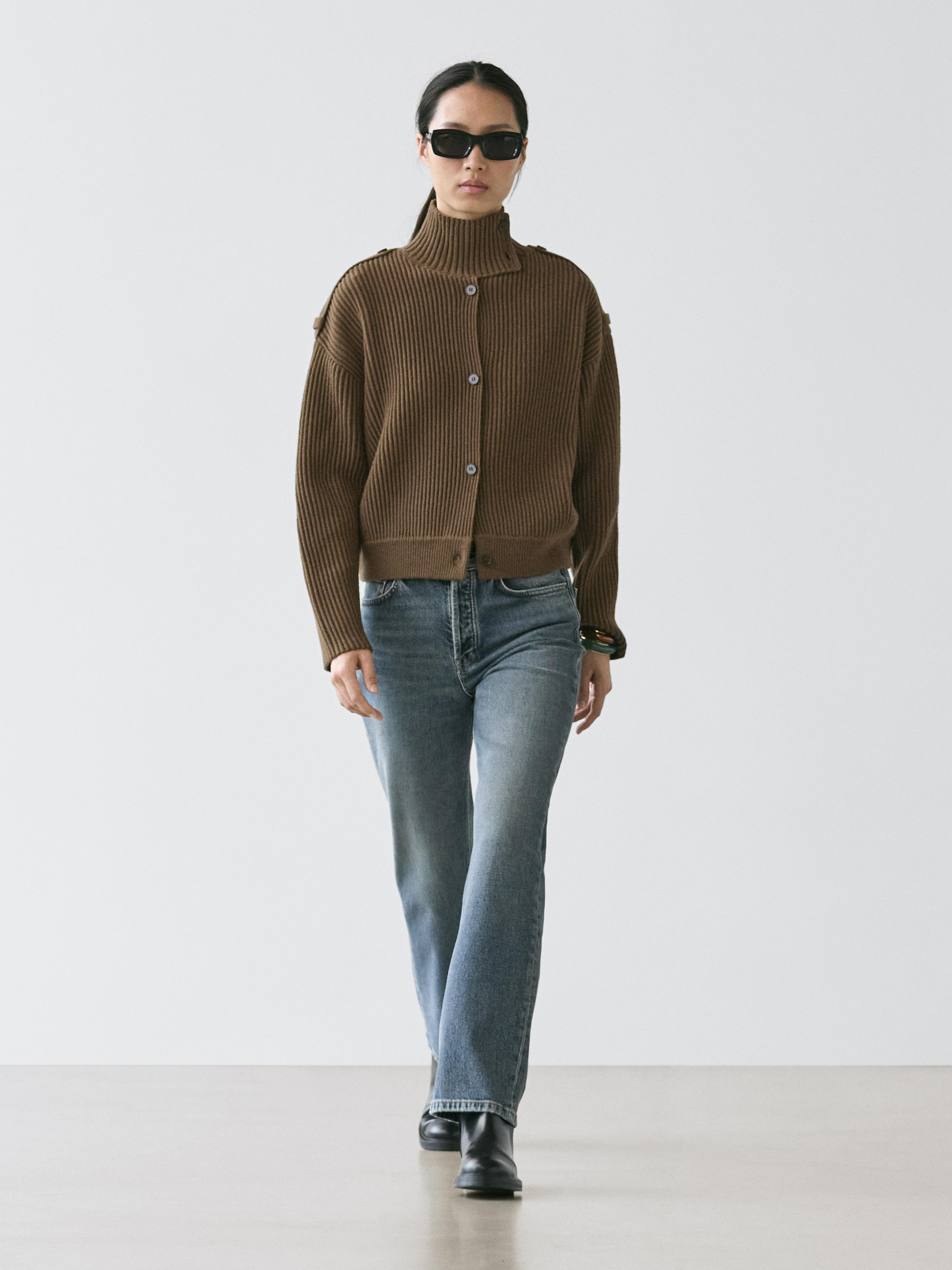 Model wears sunglasses, khaki funnel neck cardigan and blue jeans