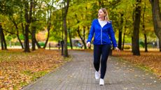 A woman in leggings, sneakers, t-shirt and a light jacket walks on a paved path. Brown leaves cover the grass either side of the path and we can see trees in the background.