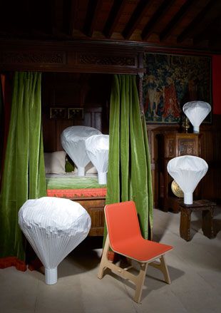 Interior bedroom, grey floor, double four poster bed, green draped curtains, wood frame, large wood headboard, white balloon shaped lamps, orange chair, wood beam ceiling, wood panelled and print walls