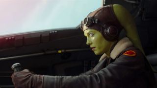Hera Syndulla is a green-skinned Twi'lek with two head tails (known as lekku) protruding from the top left and right of her head. She is wearing a brown leather cap, goggles, and a brown leather aviator jacket with a fluffy collar. Here she is piloting a spaceship.