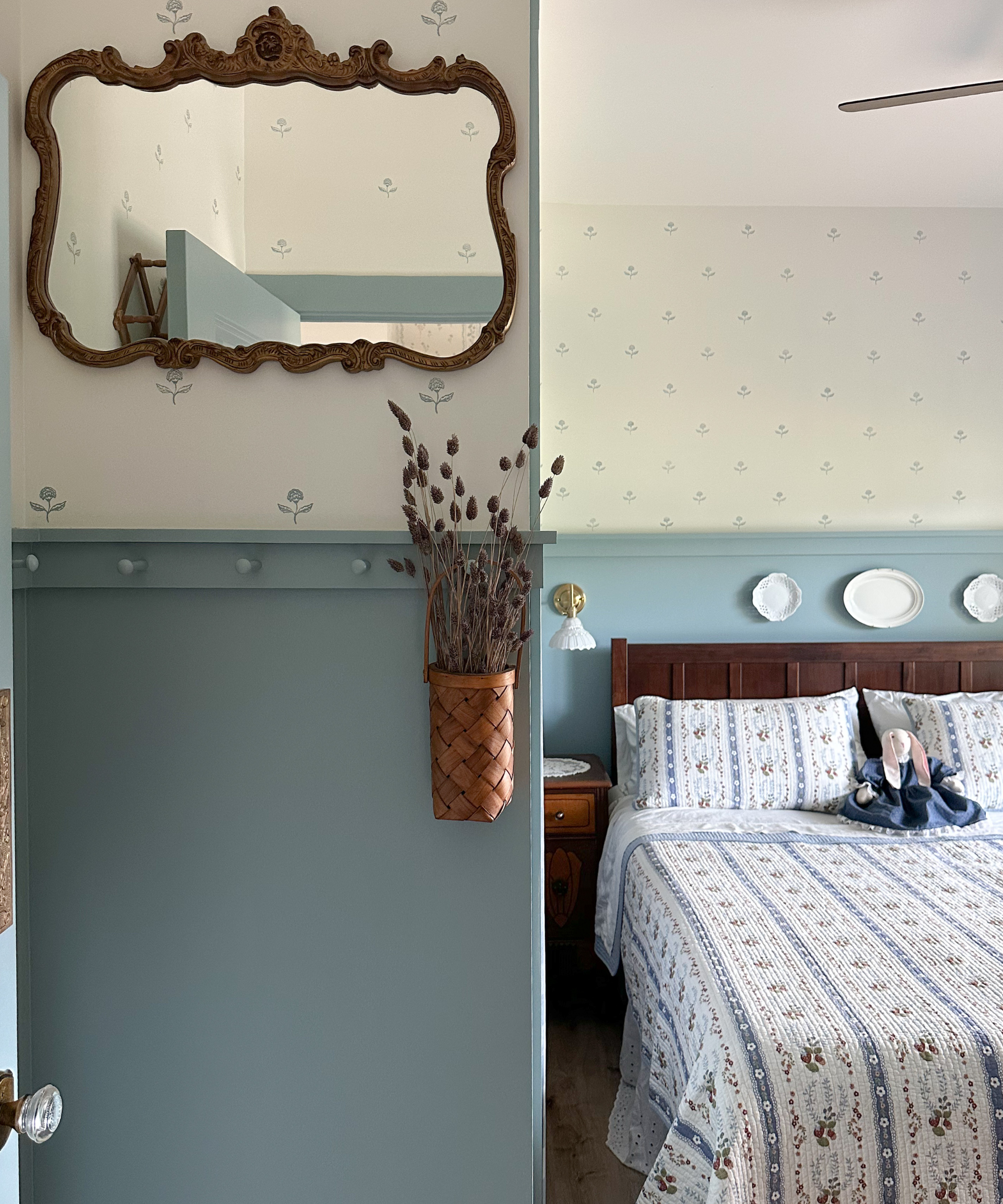 a bedroom with vintage bedding, split walls with the bottom painted blue and the top decorated with floral stamps, a basket of lavender, and a vintage mirror