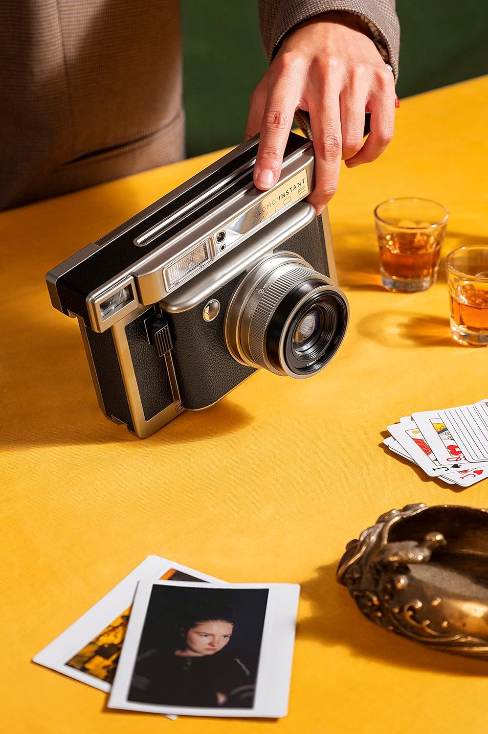 Person holding a Lomo'Instant Wide Monte Carlo on a yellow table covered with shots of alcohol, playing cards and instant photographs